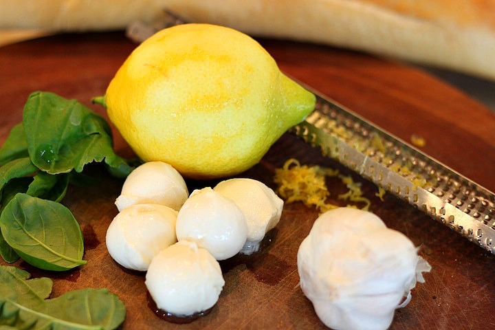 A lomon, balls of mozzarella cheese, basil leaves and a head of garlic on a wood cutting board