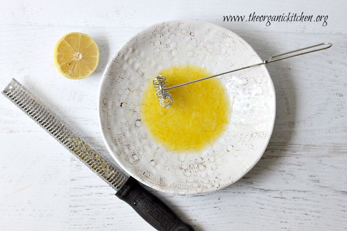 A white bowl with olive oil and lemon juice, a whisk, microplane and slice of lemon rest on a white background