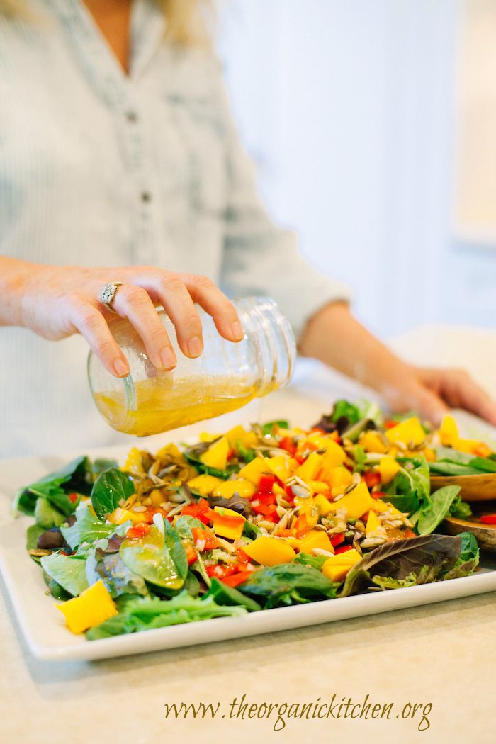 A woman pouring salad dressing on  a platter of Greens with Mango and Citrus Vinaigrette