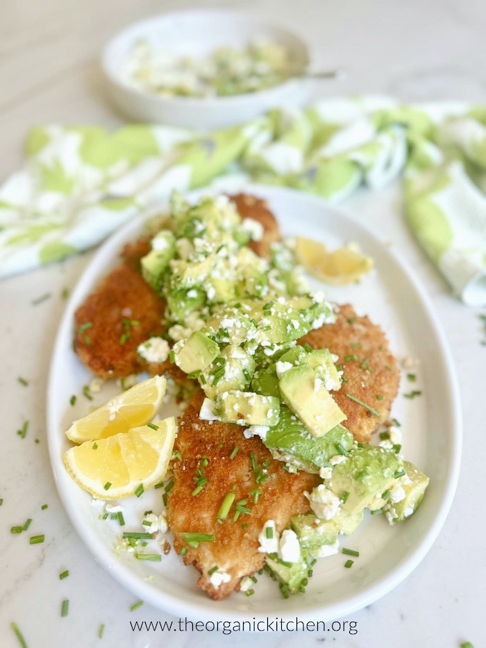 Crispy Chicken with Avocado Feta Salad on white plate with bowl of salad and dish towel in the backround