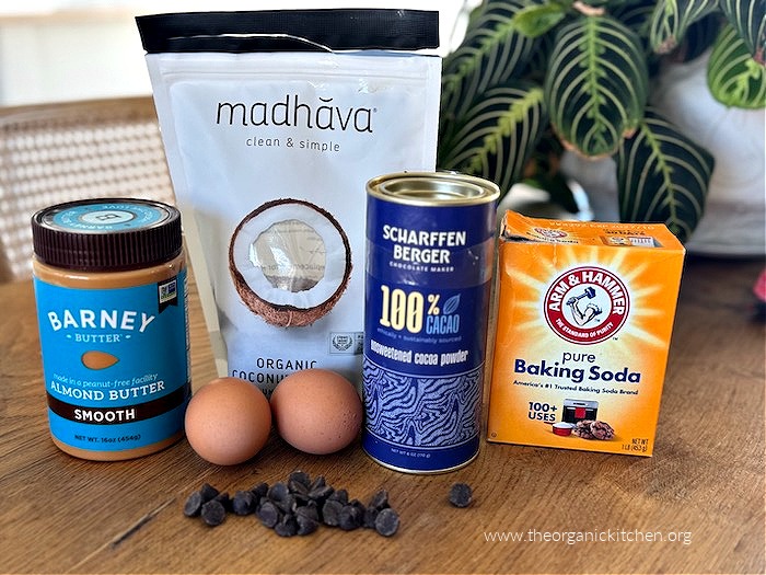 The ingredients for Healthier Brownies (High Protein-Gluten/Grain Free) sitting on a wood table: a jar of almond butter, two eggs, cocoa powder, a box of baking soda, a bag of coconut sugar and chocolate chips